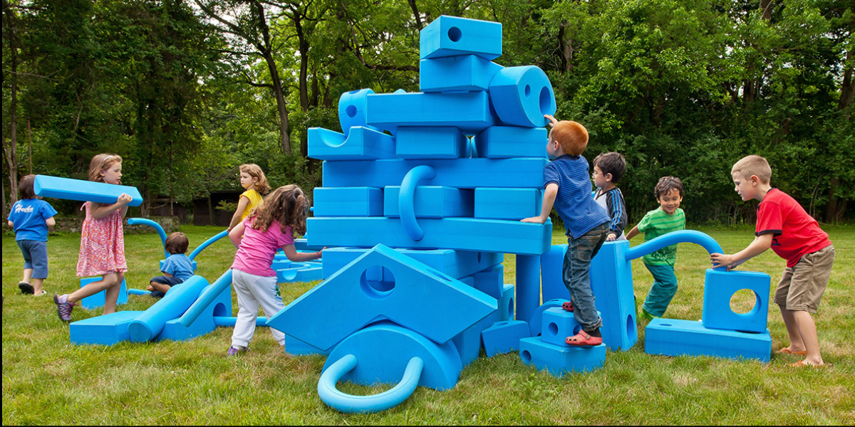 young children climb on big blue blocks in an outdoor setting. The blocks are as large as the children are.