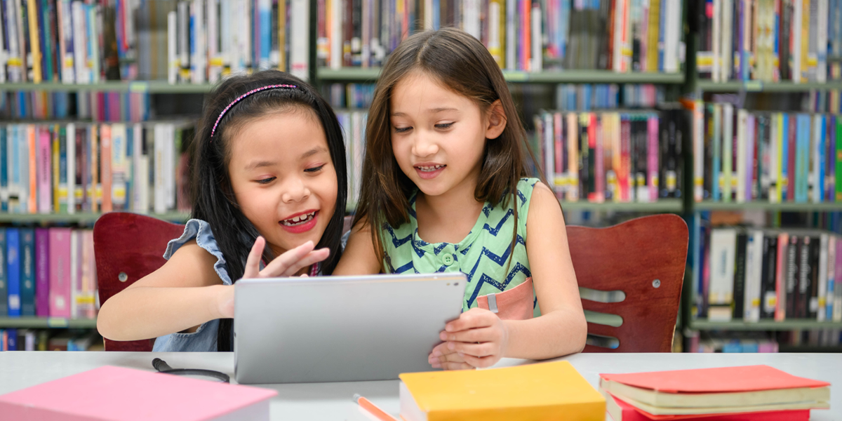 Photo of two school-aged children at a table with bookshelves in the background; the children are seated at a table where they are using a touchscreen computer or tablet