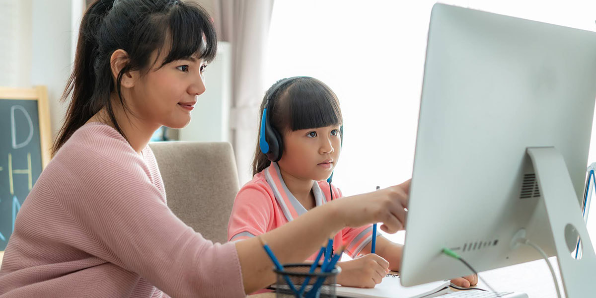 Parent and grade school child at a desk, looking at a computer screen; parent pointing, child wearing headphones