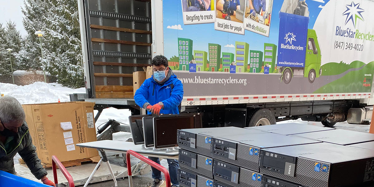 Two BlueStarRecycling workers, wearing face masks and gloves, work outside the library, readying computer components to be loaded onto the company's waiting truck, seen behind