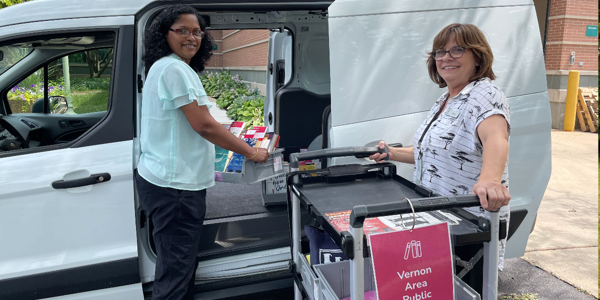 Two adult women stand next to a white van with an open door. They have a cart of library materials with them and are loading it into the van.